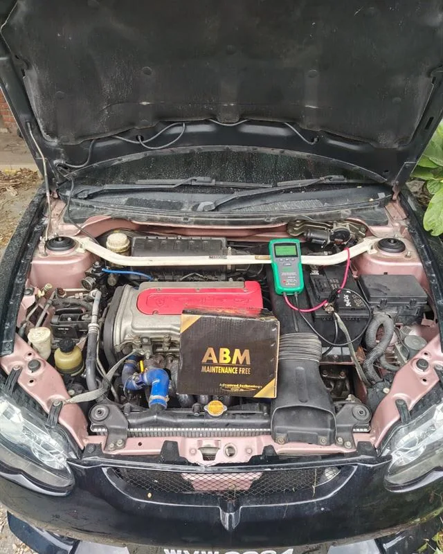 Proton Satria engine bay with a new ABM battery and a digital battery tester clipped to the terminals