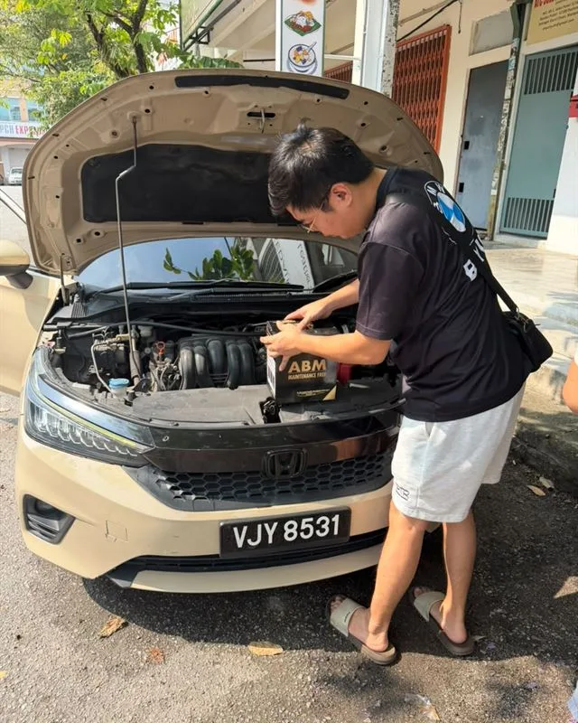 Technician lifting a new ABM Maintenance Free battery into the engine bay of a beige Honda City at the kerbside
