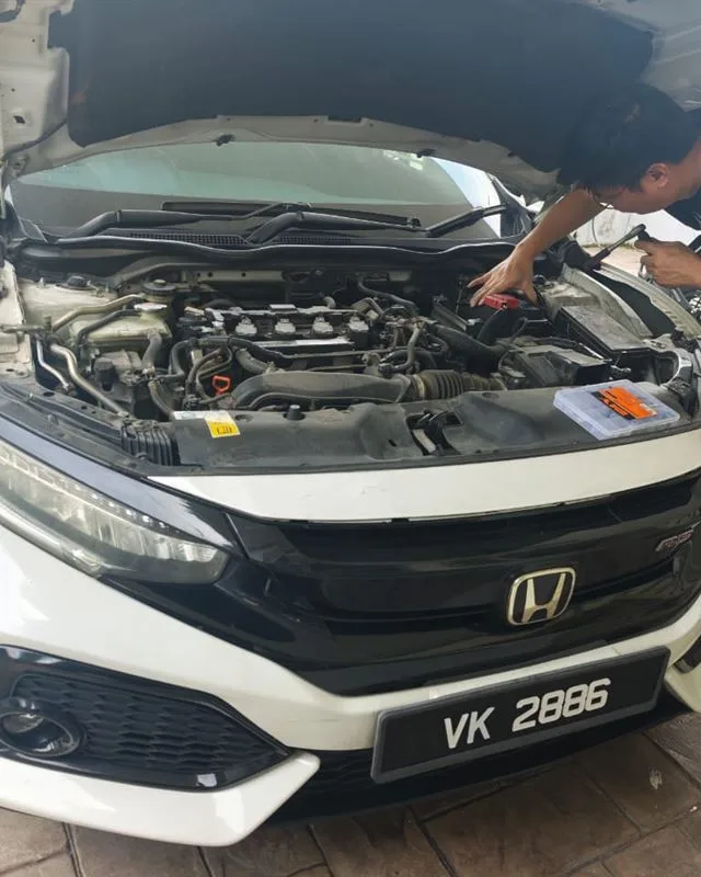 Technician leaning into the engine bay of a white Honda Civic on a home driveway, replacing the battery