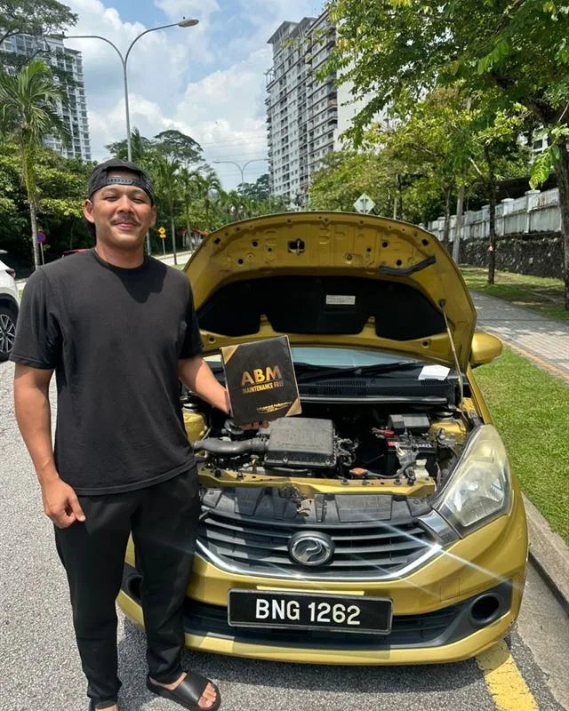 Customer standing beside a gold Perodua Myvi at the roadside holding the ABM battery box, bonnet open