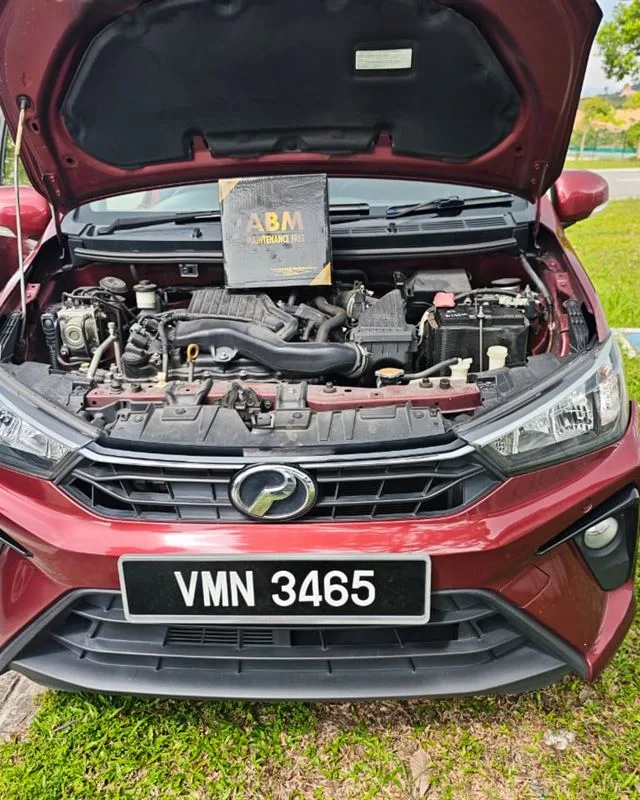 Red Perodua Bezza on a grass verge with the bonnet open and an ABM Maintenance Free battery box on the engine