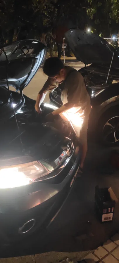 Technician working under a raised bonnet at night lit by the car headlights, toolbox on the ground beside him