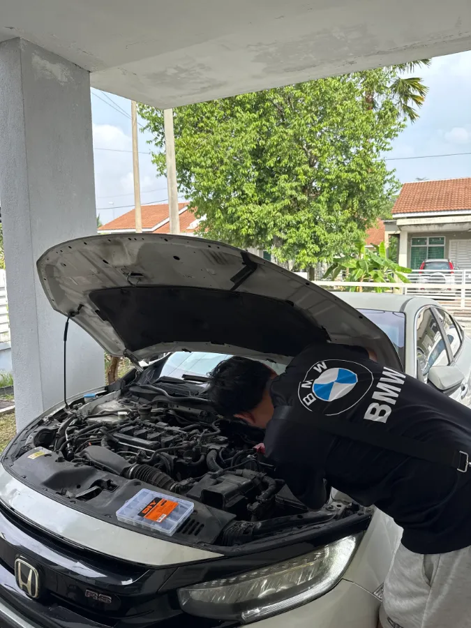 Technician leaning into the engine bay of a white Honda Civic under a carport during an on-site battery replacement